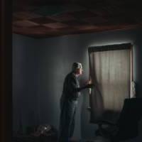 Elderly man in darkened room pulling back curtain to peer out